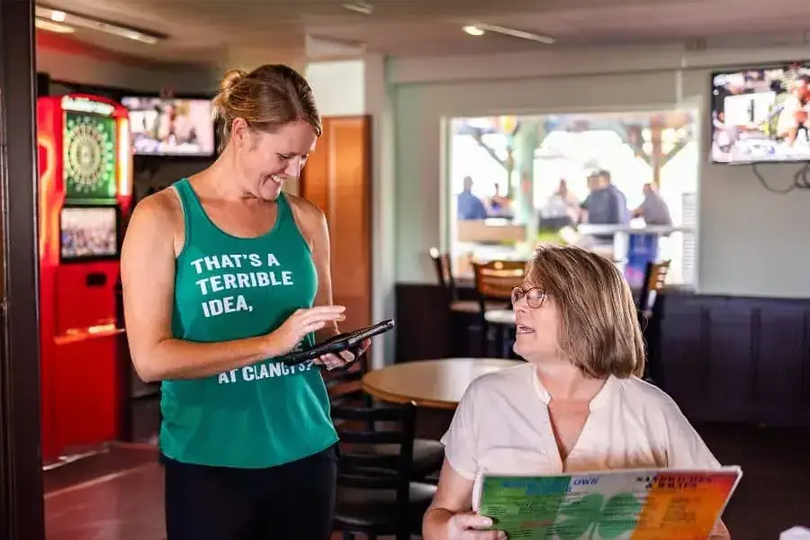 A restaurant server using a handheld POS device to take an order from a seated customer holding a menu, with a casual and friendly atmosphere in the background.