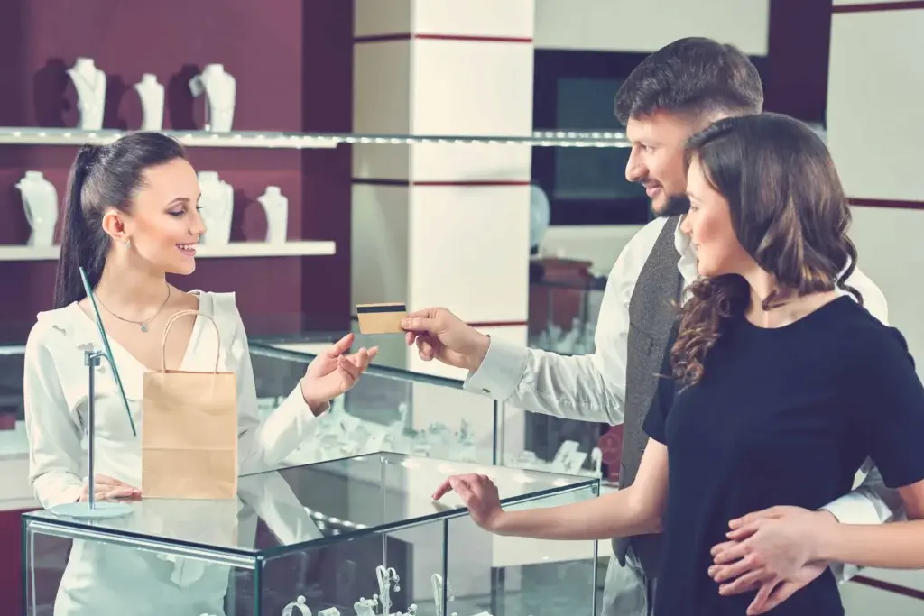 Jewelry store salesperson using a POS system to process a credit card payment from a smiling couple.