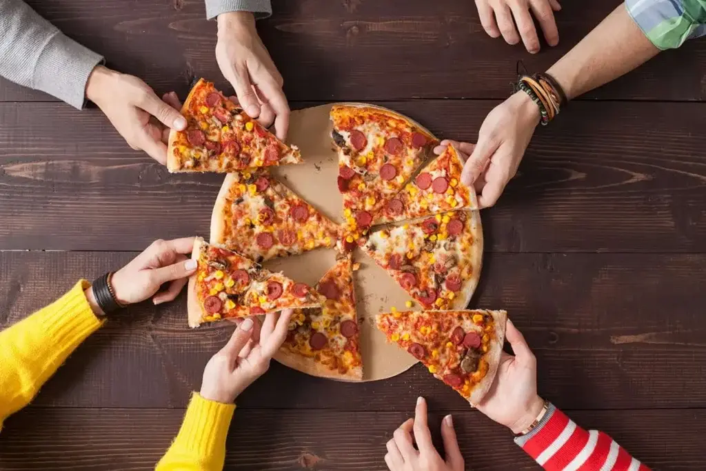 Several hands reaching for slices of a large pizza on a wooden table, symbolizing a shared dining experience in a casual pizza place.
