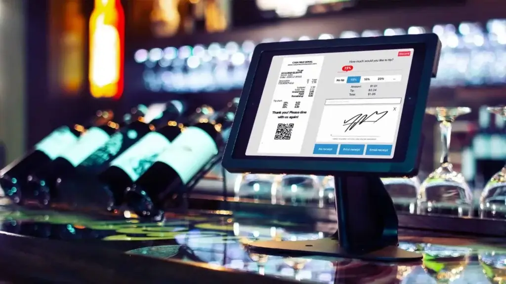A restaurant POS system displayed on a bar top, showing a digital receipt with tipping options and signature input, with bottles of wine and glasses in the background.